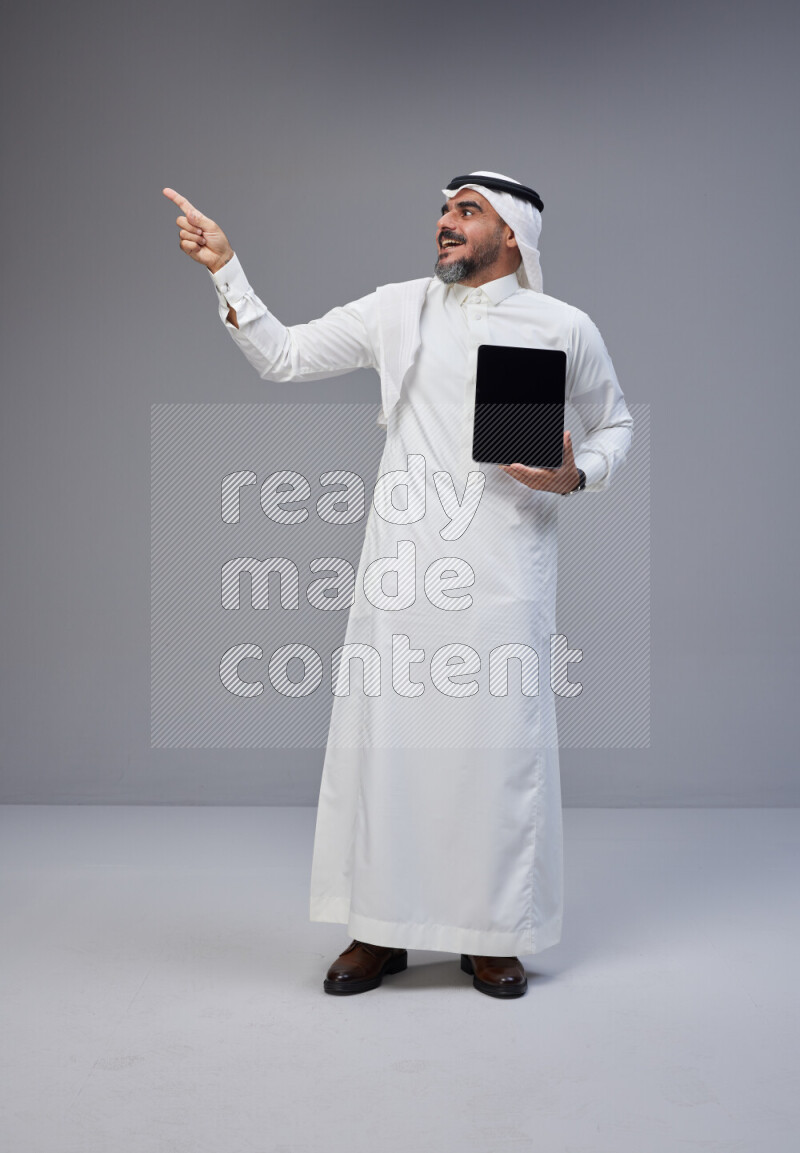 Saudi man Wearing Thob and white Shomag standing showing tablet to camera on Gray background