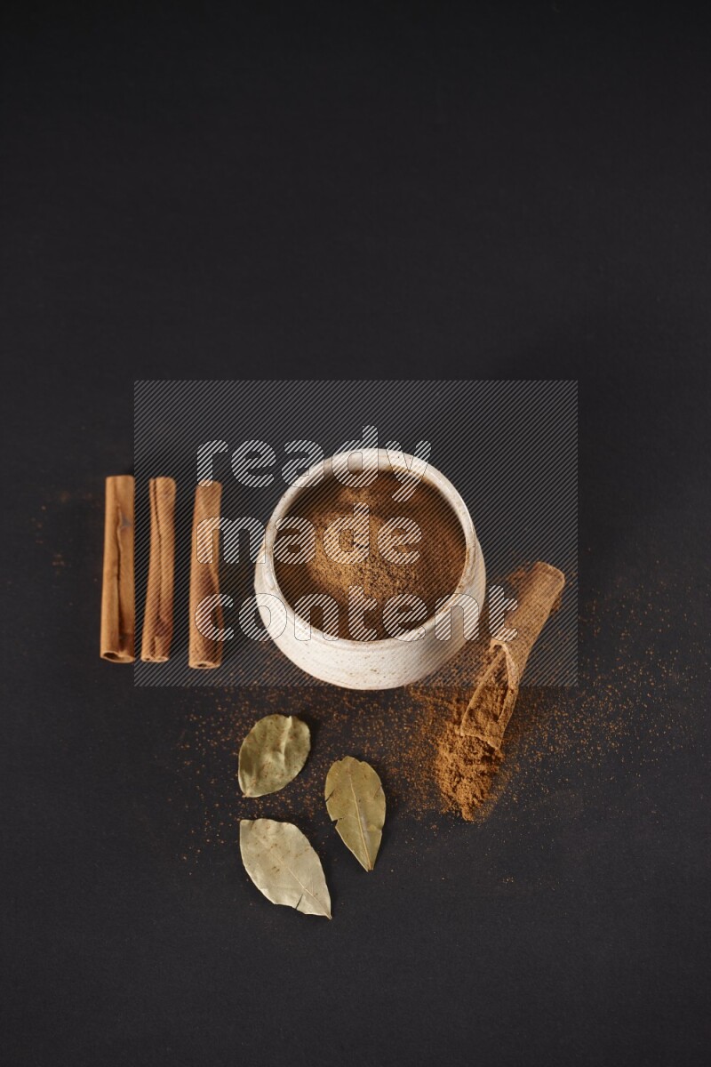 Cinnamon powder in a white pottery bowl and cinnamon sticks and laurel leaves on black background