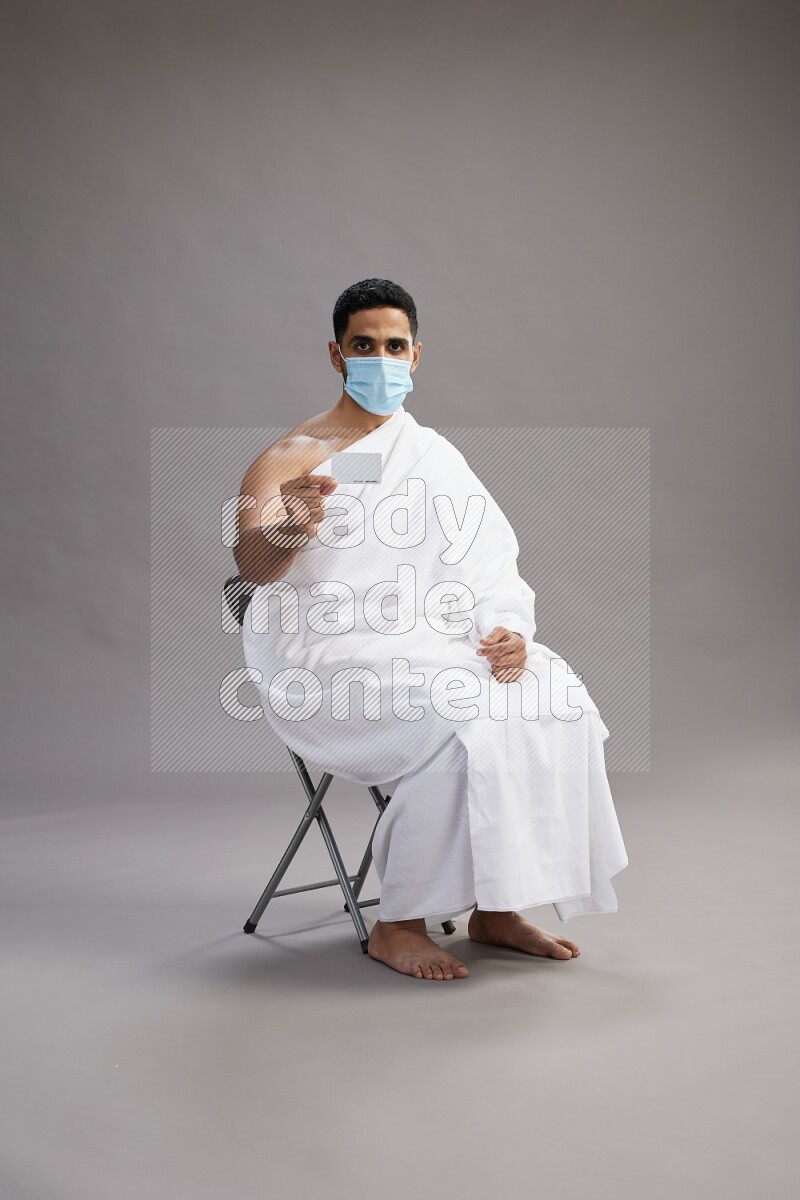 A man wearing Ehram with face mask sitting on chair holding ATM card on gray background