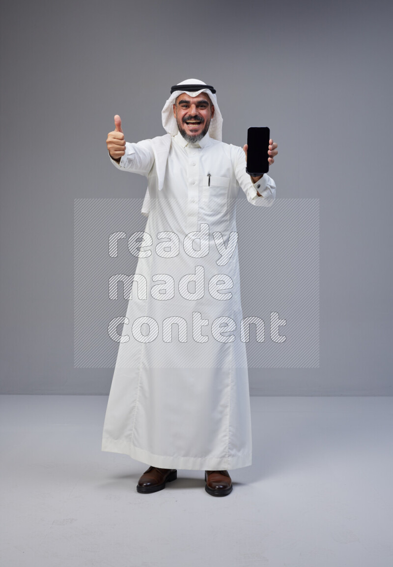 Saudi man Wearing Thob and white Shomag standing showing phone to camera on Gray background
