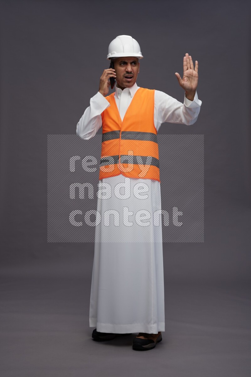 Saudi man wearing thob with engineer vest standing talking on phone on gray background