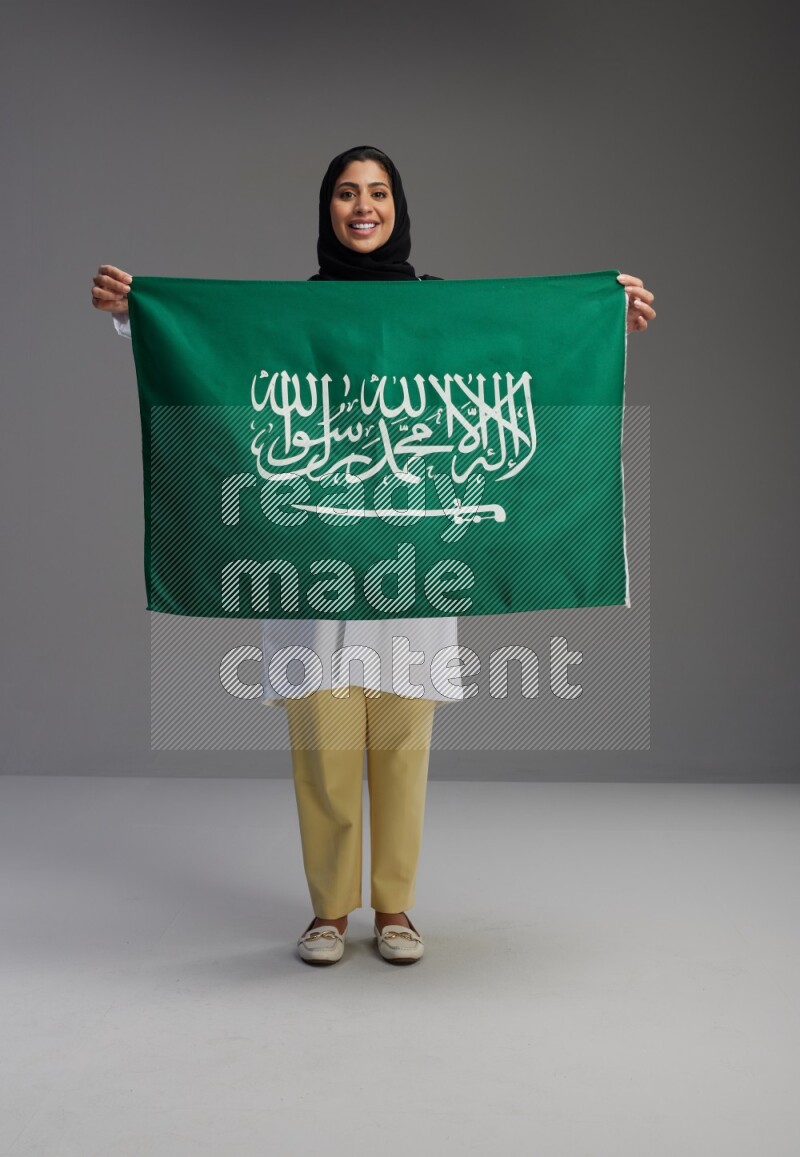 Saudi woman wearing lab coat with stethoscope standing holding Saudi flag on Gray background