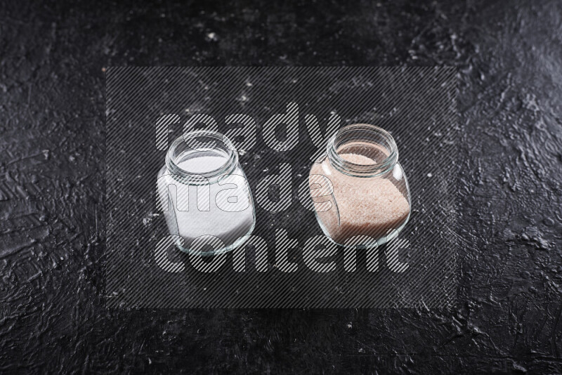 2 وعاء زجاجي احدهمها ممتلئ بملح الهيمالايا الناعم والأخر بملح الطعام على خلفية سوداء