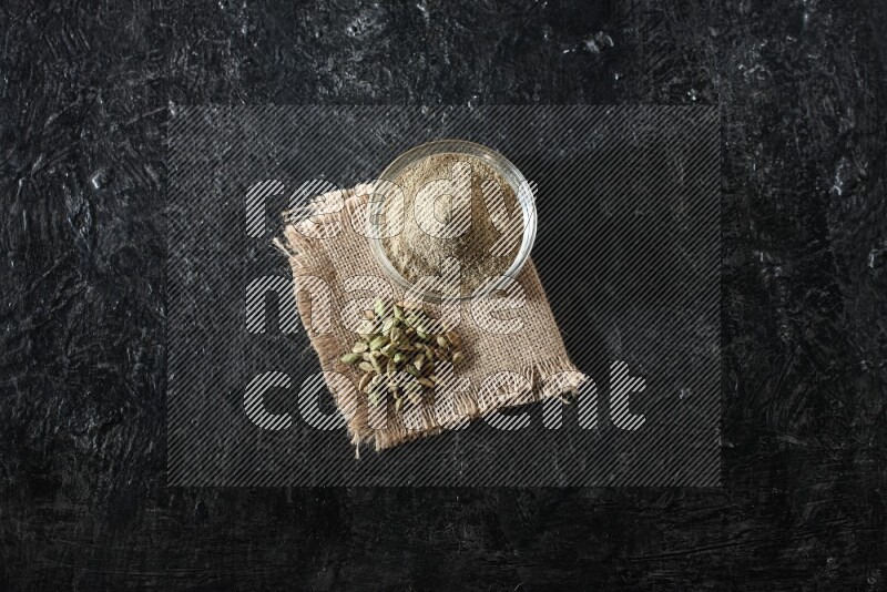 A glass bowl full of cardamom powder with cardamom seeds on a burlap piece on textured black flooring