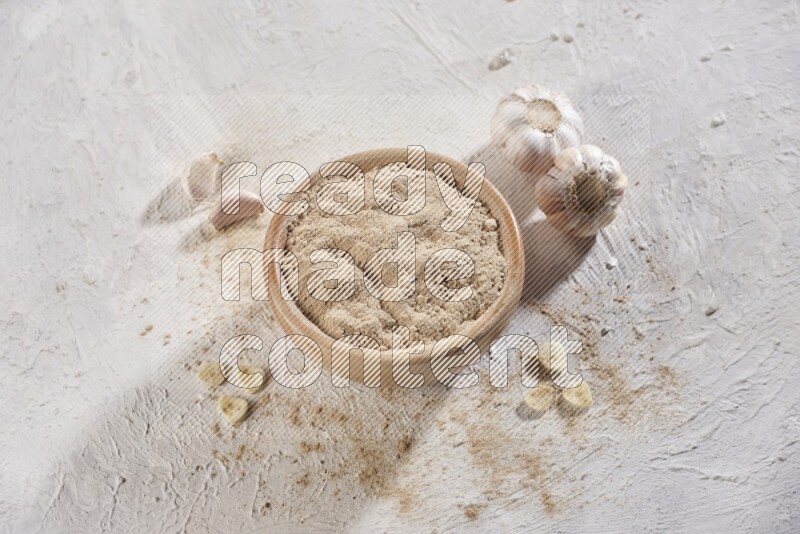 A wooden bowl full of garlic powder and beside it garlic cloves on a textured white flooring in different angles
