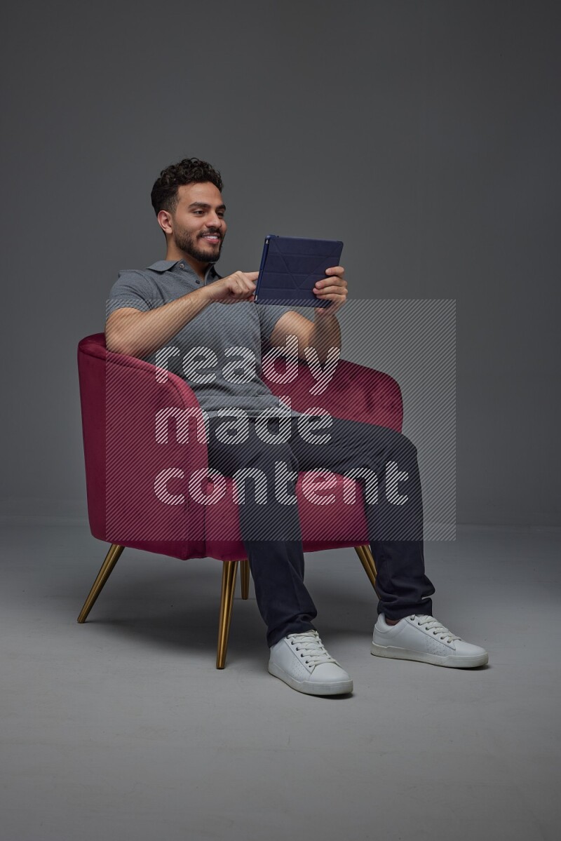A man wearing casual and using his tablet while sitting on a burgundy chair eye level on a gray background