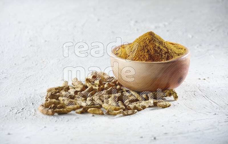 A wooden bowl full of turmeric powder and dried whole fingers beside it on a textured white flooring