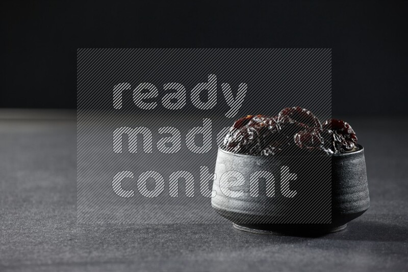 A black pottery bowl full of dried plums on a black background in different angles