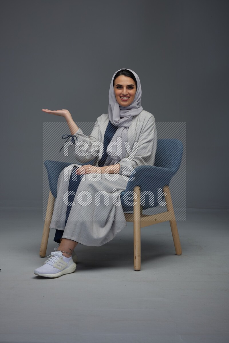 A Saudi woman wearing a light gray Abaya and head scarf sitting on a dark grey chair while making multi hand gestures eye level on a grey background