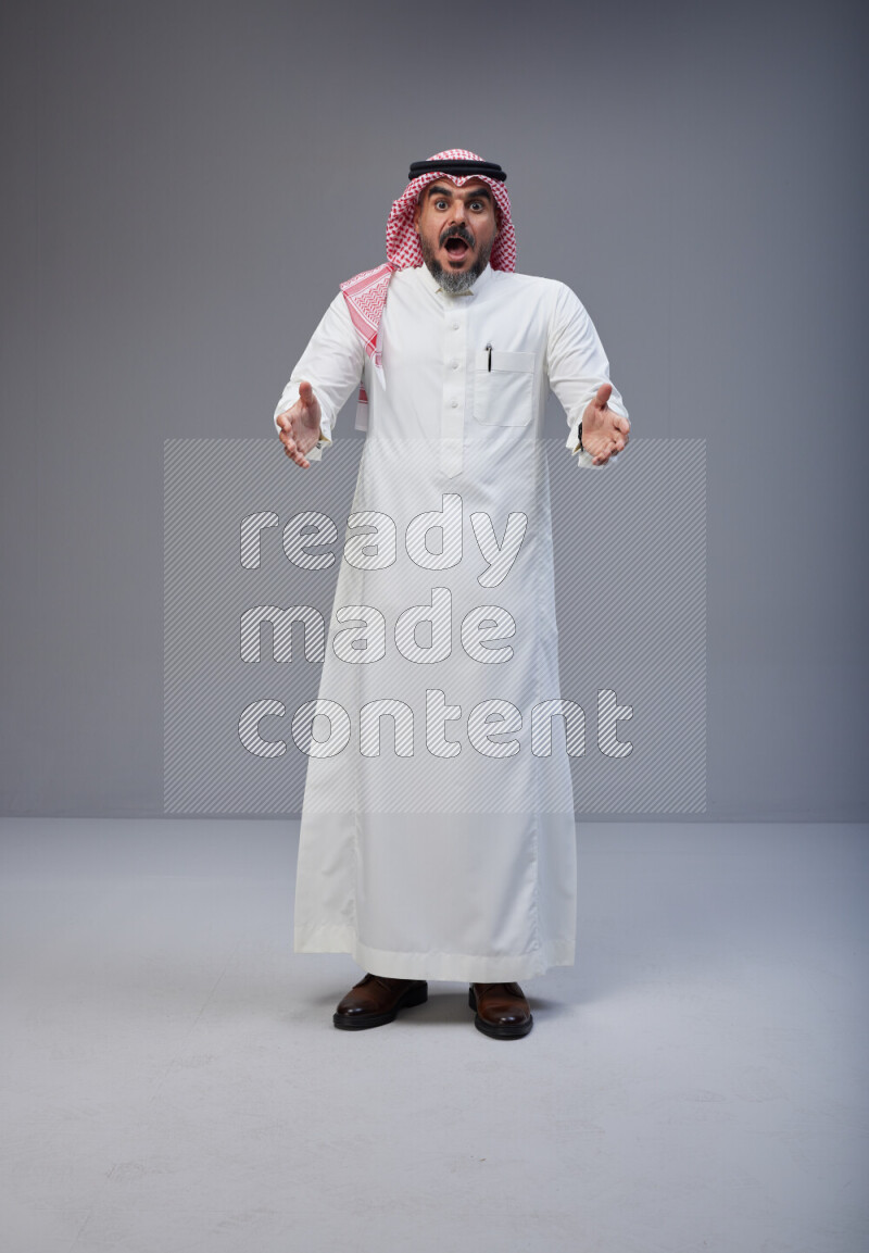 Saudi man Wearing Thob and red white Shomag standing interacting with the camera on Gray background