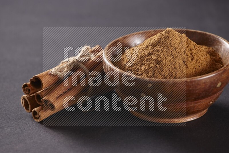 Cinnamon sticks stacked and bounded beside a wooden bowl full of cinnamon powder and a wooden spoon full of powder on black background