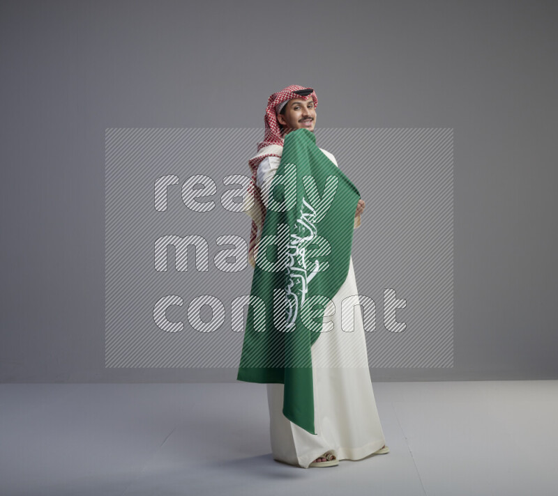 A saudi man standing wearing thob and red shomag holding big saudi flag on gray background