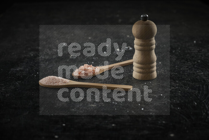 2 wooden spoons filled with fine and coarse salt with wooden grinder beside them on black background