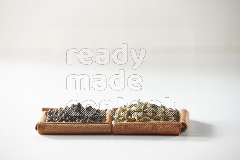 مربعان من أعواد القرفة ممتلئان بحبوب الهيل وحبوب الفلفل الأسود علي خلفية بيضاء