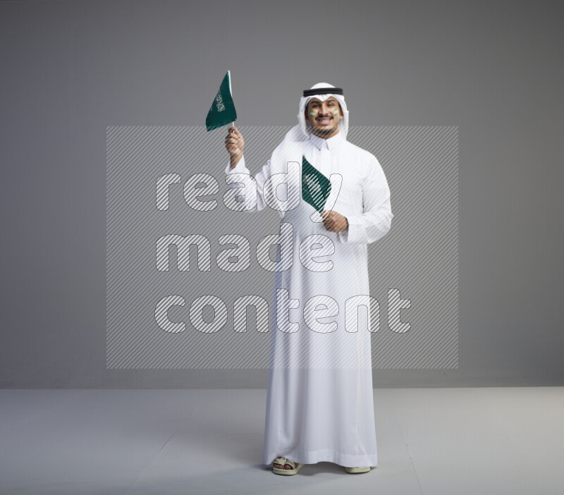 A Saudi man standing wearing thob and white shomag with face painting raising small saudi flag on gray background