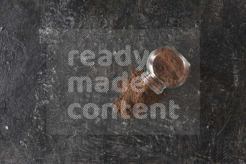 A flipped glass spice jar full of cloves powder and powder came out of it on textured black flooring