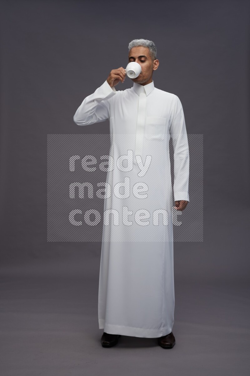 Saudi man wearing thob standing holding mug on gray background