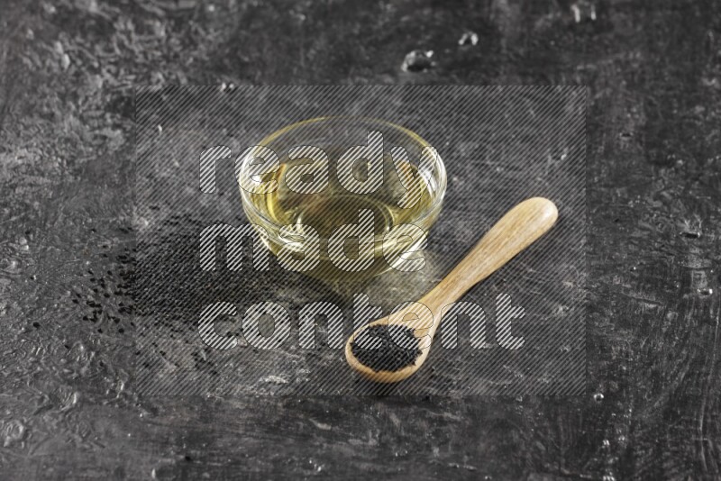 A glass bowl full of black seeds oil and wooden spoon full of black seeds with seeds spreaded on a textured black flooring