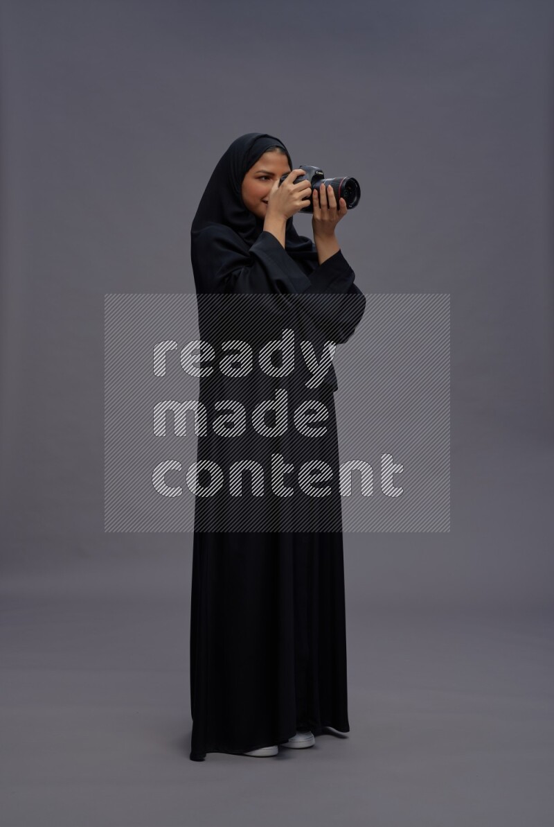 Saudi woman wearing Abaya standing holding Camera on gray background