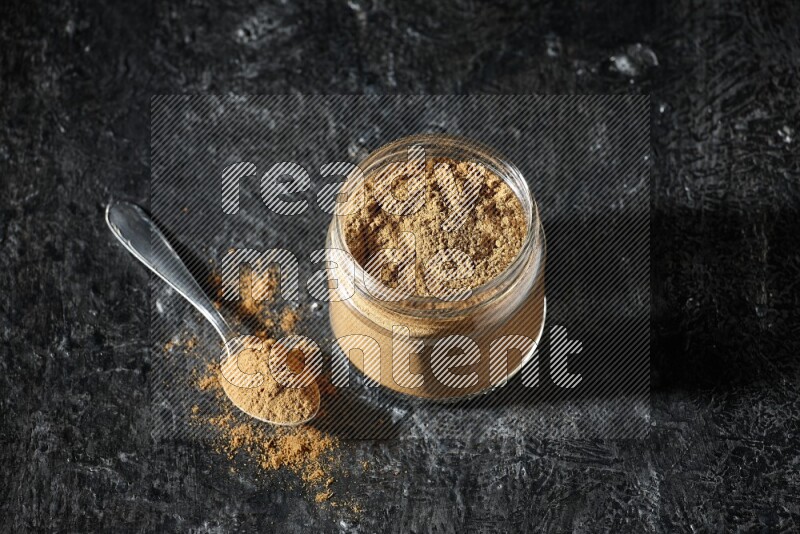 A glass jar and a metal spoon full of allspice powder on a textured black flooring