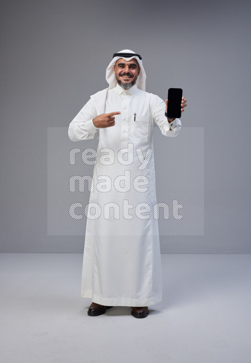 Saudi man Wearing Thob and white Shomag standing showing phone to camera on Gray background