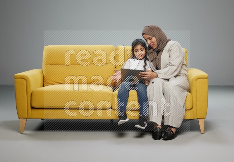 A girl with her mother on yellow sofa and watching on iPad on gray background