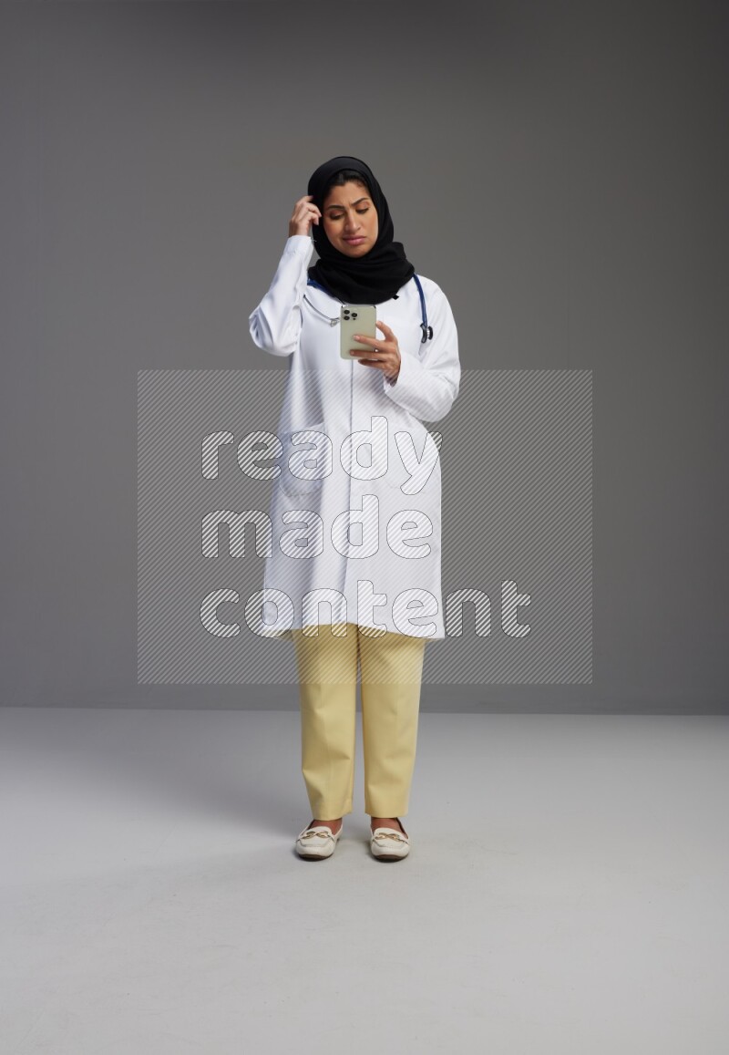 Saudi woman wearing lab coat with stethoscope standing texting on phone on Gray background