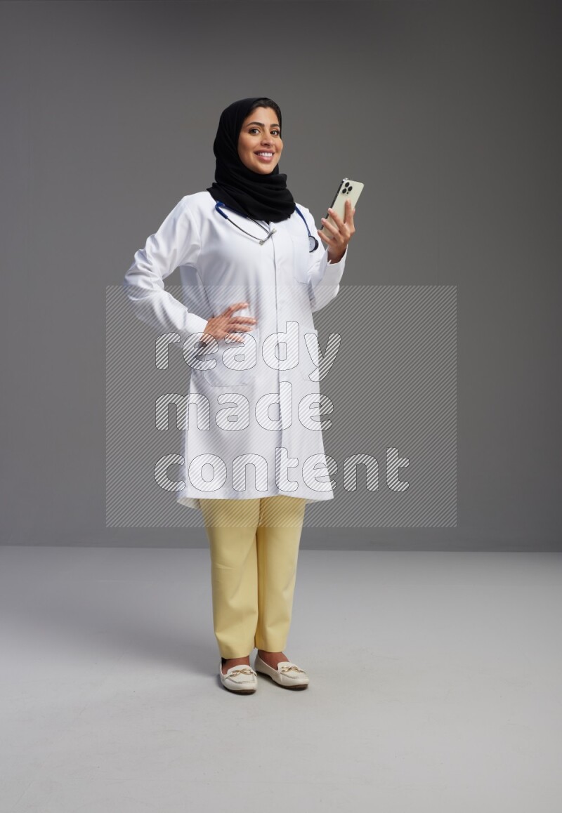Saudi woman wearing lab coat with stethoscope standing texting on phone on Gray background