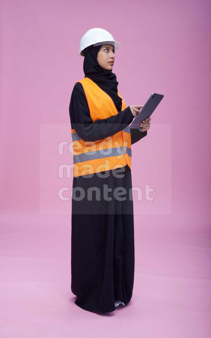 Saudi woman wearing Abaya with engineer vest and helmet standing working on tablet on pink background