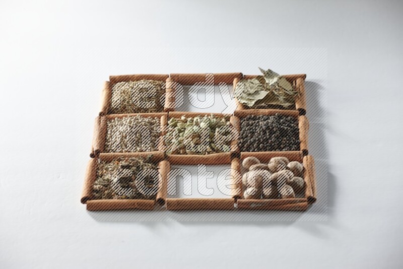 9 مربعات من أعواد القرفة ممتلئة بالهيل في المنتصف محاطة بجوزة الطيب والملح والريحان المجفف والفلفل الأبيض والكمون وبهار الفلفل الحلو وأوراق الغار والفلفل الأسود على أرضية بيضاء