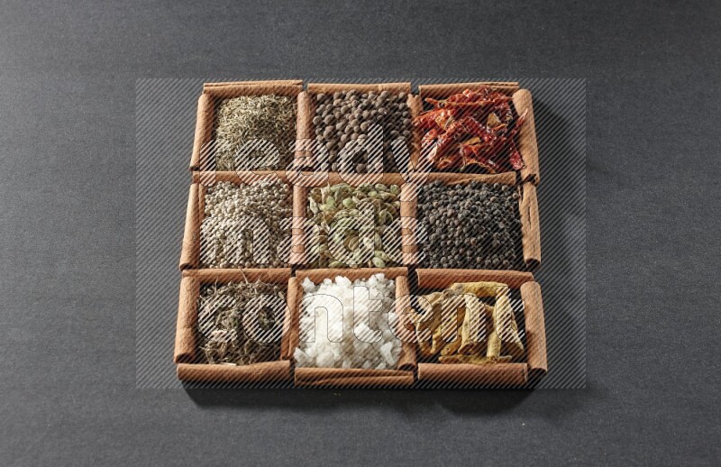 9 squares of cinnamon sticks full of cardamom in the middle surrounded by turmeric fingers, black pepper, chilis, allspice, cumin, white pepper, dried basil and salt on black flooring