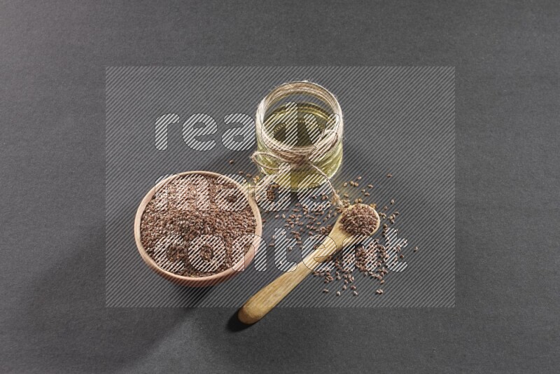 A wooden bowl and wooden spoon full of flaxseeds with a glass jar of flaxseeds oil on a black flooring