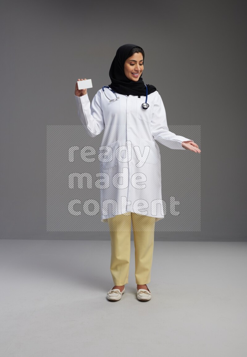 Saudi woman wearing lab coat with stethoscope standing holding ATM card on Gray background