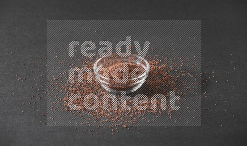 A glass bowl full of garden cress seeds surrounded by seeds on a black flooring