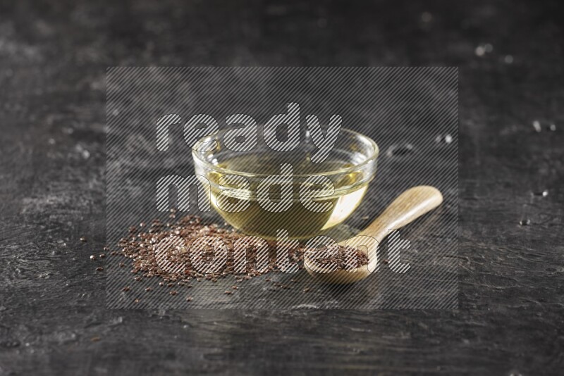 A glass bowl full of flaxseeds oil and wooden spoon full of flaxseeds with seeds spread on a textured black flooring