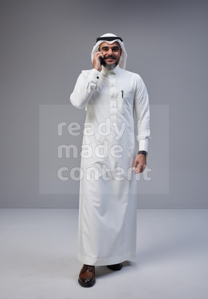 Saudi man Wearing Thob and white Shomag standing talking on phone on Gray background