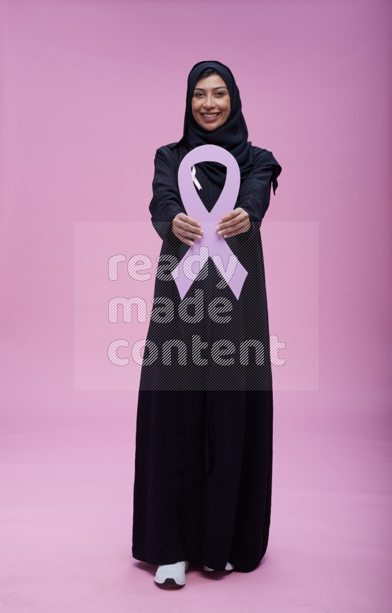 Saudi woman wearing pink ribbon on abaya standing holding awareness ribbon on plain pink background