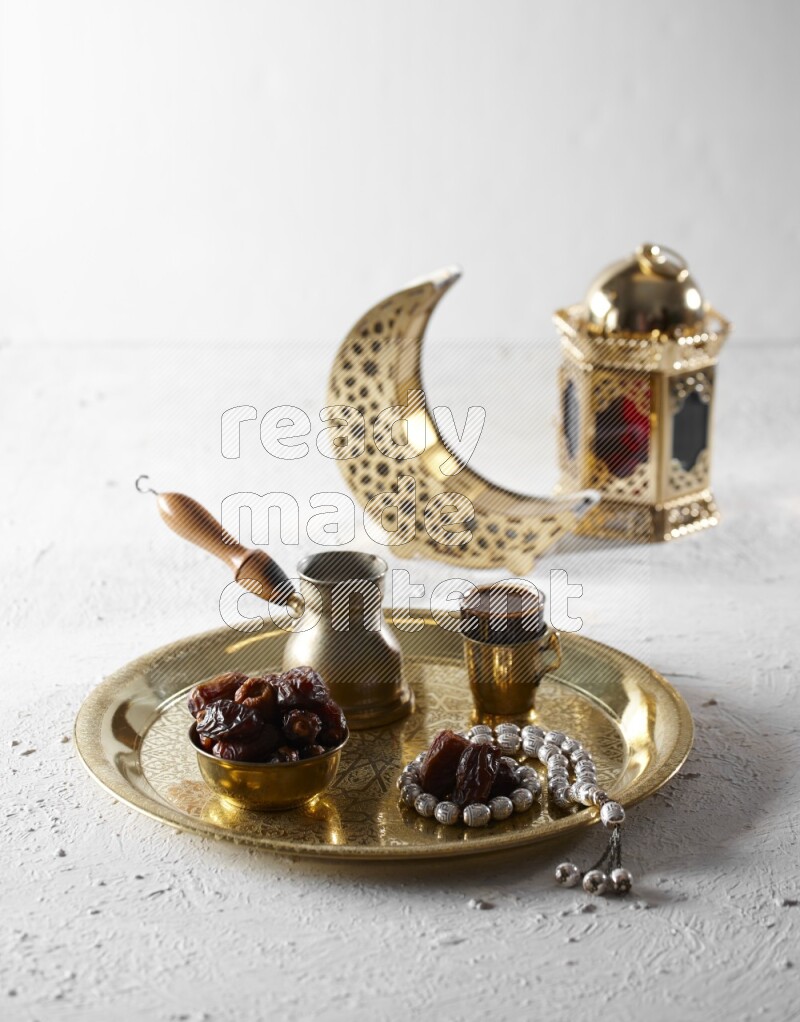 Dates in a metal bowl with coffee and prayer beads on a tray beside lanterns in a light setup