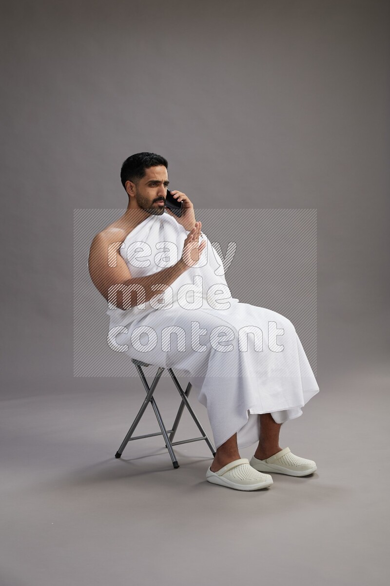 A man wearing Ehram sitting on chair talking on the phone on gray background