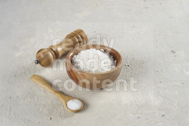 A wooden bowl and spoon filled with white sea salt and wooden grinder beside them on white background