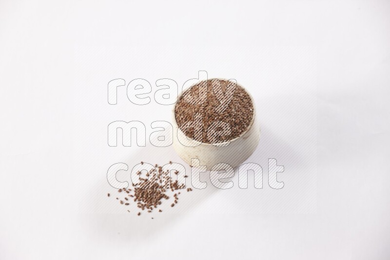 A pottery beige bowl full of flax seeds and more seeds spread on a white flooring