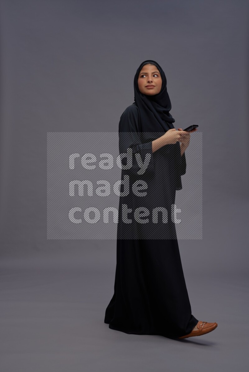 Saudi woman wearing Abaya standing texting on phone on gray background