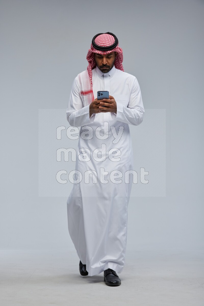 Saudi man Wearing Thob and shomag standing texting on phone on Gray background