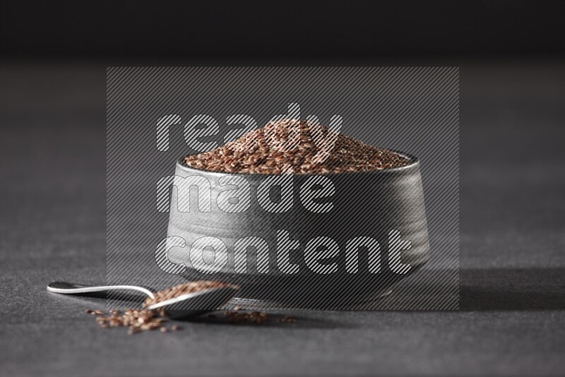 A black pottery bowl full of flaxseeds and a metal spoon full of the seeds on a black flooring