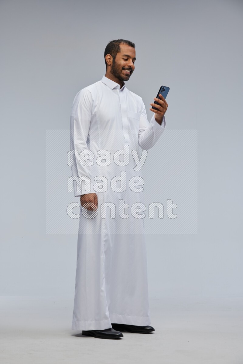 Saudi man wearing Thob standing texting on phone on Gray background