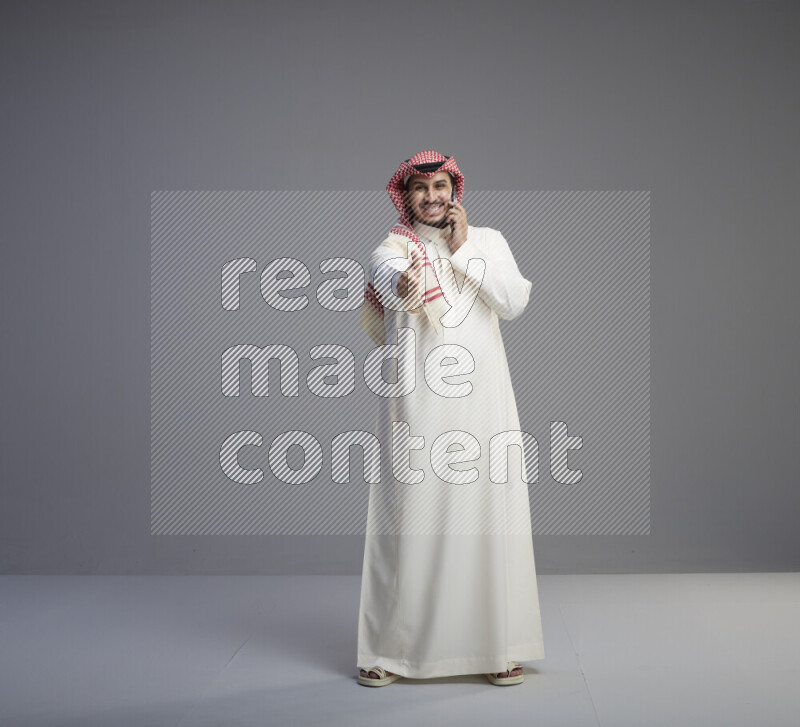 A Saudi man standing wearing thob and red shomag talking on phone on gray background