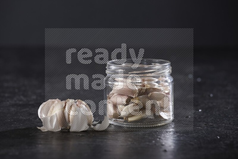 A glass jar full of garlic cloves with a whole garlic bulb beside it on a textured black flooring