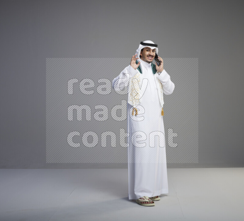 A saudi man standing wearing thob and white shomag with flag scarf on his neck talking on phone on gray background