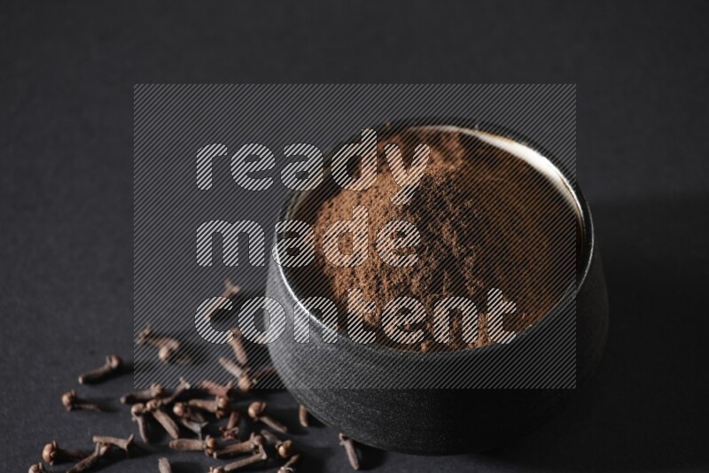 وعاء فخاري أسود ممتلئ ببودرة القرنفل وحبوب القرنفل على خلفية سوداء