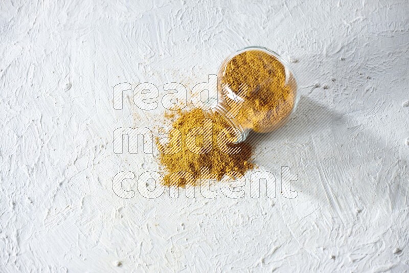 A flipped glass spice jar full of turmeric powder and powder fell out it on textured white flooring